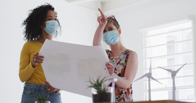 The image shows two women wearing face masks and discussing a blueprint while pointing towards something, indicating teamwork and collaboration. Suitable for use in topics related to architecture, urban planning, COVID-19 safety protocols, and office teamwork. The presence of wind turbine models could also suggest discussions related to sustainable energy or renewable sources.