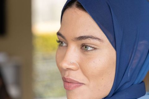 Young woman wearing blue hijab gazing out window with nose stud and natural makeup