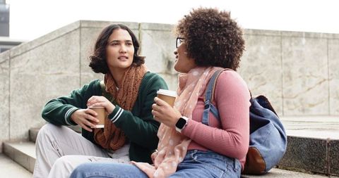 Multiethnic young women chatting with to-go coffee on urban stone steps, autumn lifestyle