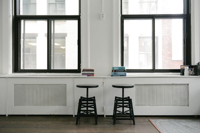 Minimalist workspace features two black stools placed in front of large windows, with multiple books stacked on adjacent surfaces. Suitable for illustrating modern office environments, creative studio setups, or spacious work areas.