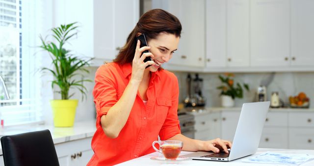 Smiling woman working at home kitchen with laptop and mobile phone - Download Free Stock Images Pikwizard.com