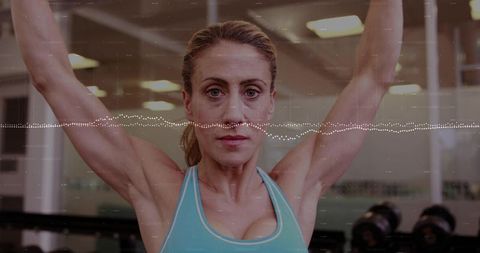 Focused mid-adult woman raising arms overhead in gym wearing turquoise sports bra