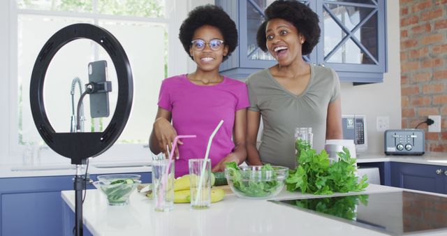 This image depicts a mother and daughter actively engaged in their kitchen while recording a cooking video with a ring light and smartphone. The scene is filled with fresh vegetables and fruits, suggesting health and wellness themes. Ideal for use in promoting cooking channels, healthy lifestyle blogs, family activity features, or content about home learning and vlogging.