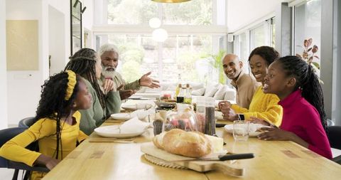 Multigenerational family passing bread around sunlit open-plan dining table, sharing meal