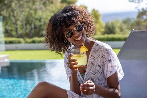 Smiling woman relaxing poolside with citrus cocktail, sunglasses and crochet cover-up