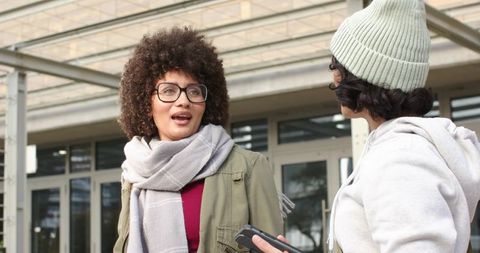 Diverse women talking at university entrance holding smartphone wearing eyeglasses