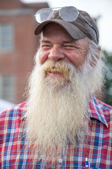 Elderly man with a long grey beard and glasses perched on his cap is smiling outdoors. He is wearing a red plaid shirt and appears cheerful and content. Could be used for themes of happiness, aging, lifestyle, or outdoors.