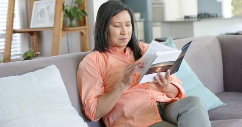 Senior asian woman reading booklet on gray sofa in sunlit living room wearing salmon shirt