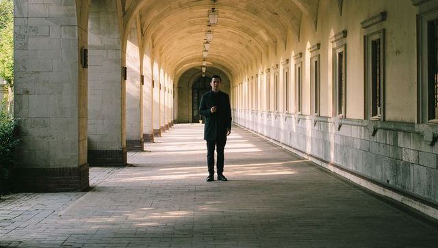 Man standing in sunlit stone colonnade holding smartphone, symmetrical arched corridor