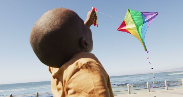 Child enjoying flying a multicolored kite at the beach with the ocean in the background. Great for topics related to outdoor activities, childhood fun, summer vacations, and marine recreation.