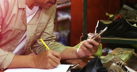 Cobbler Using Mobile Phone While Taking Notes in Workshop