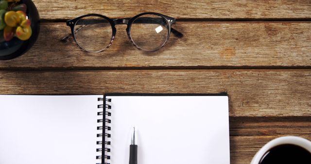 A pair of glasses rests on a wooden table next to an open notebook and a pen, with copy space. A cup of coffee and a small potted plant add a cozy atmosphere to a potential work or study environment.
