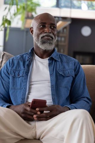 Senior african man relaxing on sofa holding smartphone looking thoughtful and confident