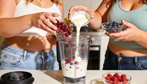 Women pouring milk and berries into blender making berry yogurt smoothie in home kitchen