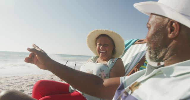 Elderly African American couple enjoying a sunny day on the beach. They are smiling and comfortable in beach chairs, wearing hats for sun protection. Ideal for use in travel advertisements, retirement planning promotions, or showcasing active senior lifestyle.