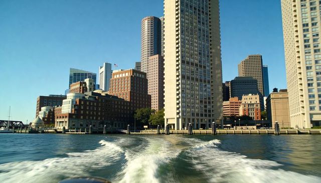 Boat wake leading to waterfront skyline with tall beige tower and harbor pilings