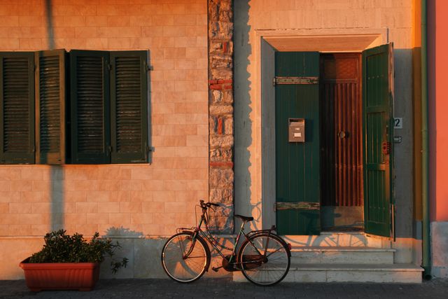 An old-fashioned bicycle leans against a house exterior with green shutters and a wooden door illuminated by warm afternoon light. Perfect for uses related to travel, Mediterranean lifestyle, vintage themes, and exterior decor.