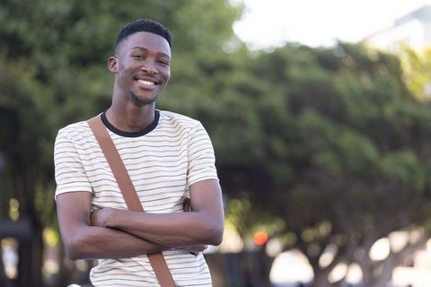 Young man standing smiling with arms crossed wearing striped shirt and crossbody bag