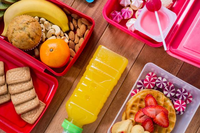 Colorful lunch boxes filled with a variety of snacks and sweets, including fruits, nuts, biscuits, juice, waffles with strawberries, and candy, arranged on a wooden table. Ideal for illustrating healthy eating, meal prep, school lunches, and snack ideas.
