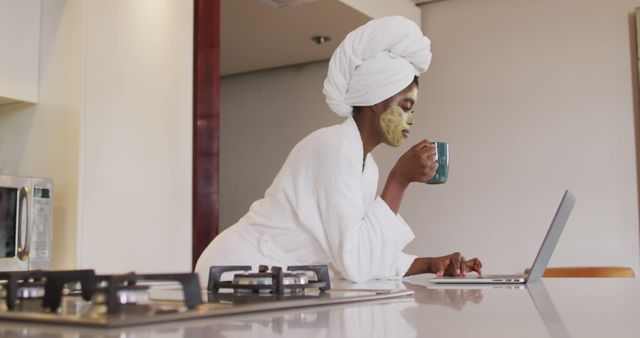 African american attractive woman with applied face mask drinking coffee and using laptop in kitchen. beauty, pampering, home spa and wellbeing concept.