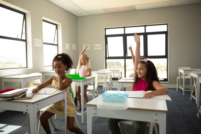 This image shows happy multiracial elementary schoolgirls raising their hands while sitting at desks in a classroom. It is ideal for use in educational materials, school brochures, websites, and advertisements promoting diversity, childhood education, and student engagement.