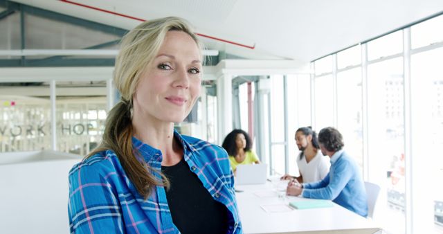 Professional woman standing confidently in foreground with her diverse business team having a discussion at a table behind her in a modern office. Useful for depicting leadership, teamwork, business environments, corporate culture, and gender diversity in the workplace visuals.