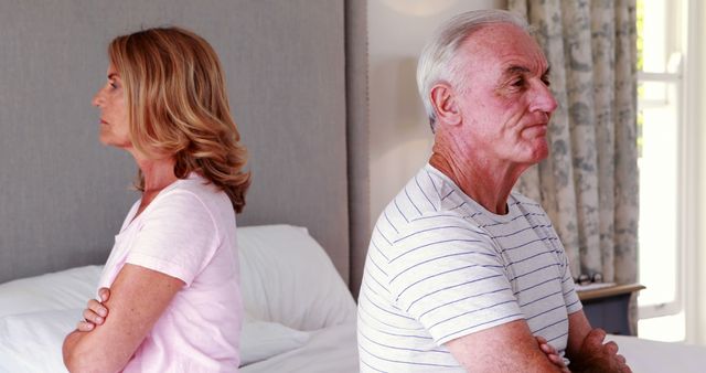 This image depicts a senior couple sitting on a bed with their backs to each other, illustrating estrangement and conflict. Their serious expressions and crossed arms signify anger and unresolved issues. Useful for articles on marital problems among older adults, relationship counseling for seniors, or illustrating emotional distance and communication issues in aging relationships.