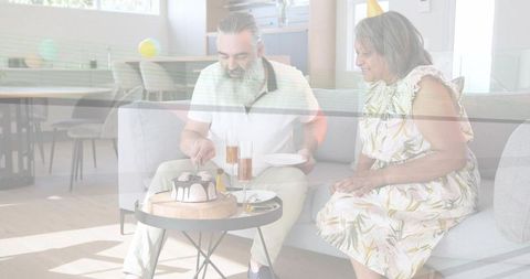 Mature couple cutting cake on coffee table at home celebrating with champagne