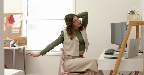 Woman stretching at home office desk by window with laptop and monitor, minimalist neutral