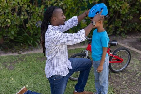 African american father fitting blue bicycle helmet on son near red bike in backyard