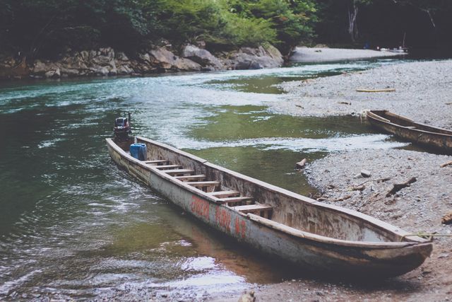 Wooden boat resting by riverbank with clear water in lush forest. Suitable for nature-themed projects, travel blogs, adventure magazines, or posters.