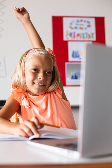 Smiling caucasian elementary schoolgirl raising hand during online class at desk in classroom. unaltered, education, intelligence, happiness, wireless technology, studying and school concept.