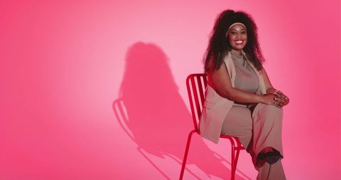 Smiling african-american woman sitting on red chair against pastel pink backdrop