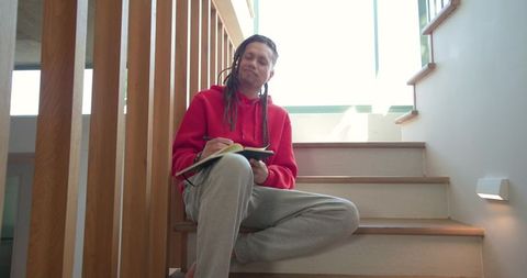 Mid-adult man writing journal on staircase in red hoodie, minimalist home interior