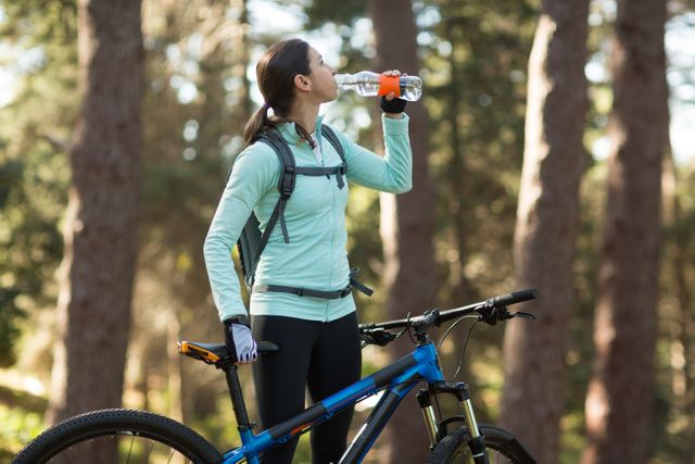 Female cyclist taking a break to drink water in a forest. Ideal for use in content related to outdoor activities, fitness, hydration, and healthy lifestyles. Can be used in advertisements for sports equipment, hydration products, or outdoor adventure promotions.