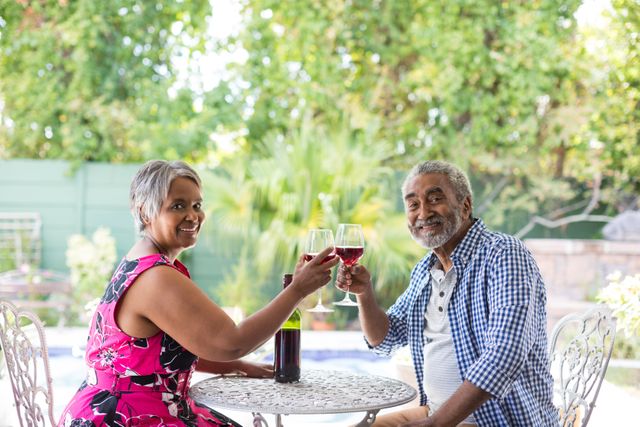 Senior couple sitting at a table in their backyard, toasting wineglasses and smiling. Ideal for use in advertisements or articles about senior lifestyle, retirement, outdoor activities, and celebrating special moments together.