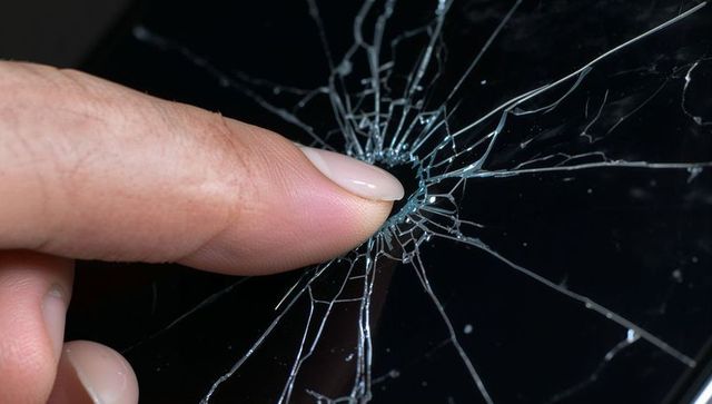 Manicured fingertip pressing into glass causing radial spiderweb crack with micro shards