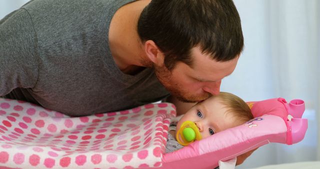 Caucasian man gently kisses baby at home. Tender father-daughter moment captured in a cozy domestic setting.