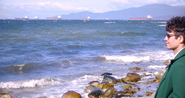 Man standing on rocky shore overlooking vast ocean with several ships on horizon. Ideal for themes of contemplation, travel, nature, and peaceful scenery. Useful in travel magazines, nature blogs, or mental well-being articles focused on relaxation and mindfulness.