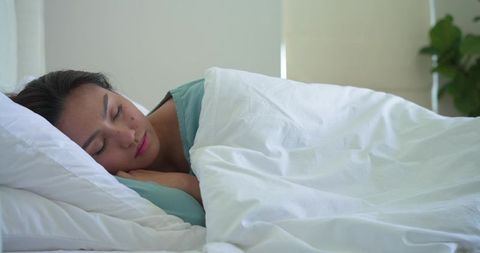 Asian woman sleeping peacefully under white duvet in daylight bedroom wearing teal top