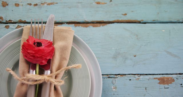 A neatly presented table setting features a fork and knife wrapped in a beige napkin, adorned with a red flower, on a rustic blue wooden table, with copy space. It evokes a sense of homely elegance, ideal for a casual yet stylish dining experience.