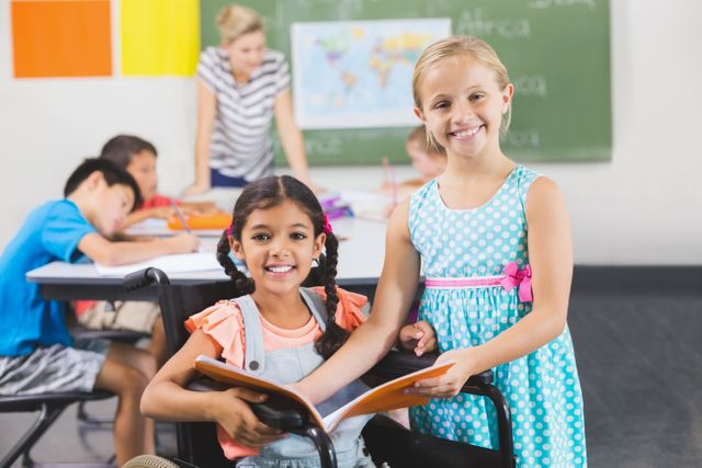 Two schoolgirls, one in a wheelchair, smiling and holding a book in a classroom. Other children and a teacher are in the background. Ideal for educational materials, promoting inclusivity, and teamwork in schools.