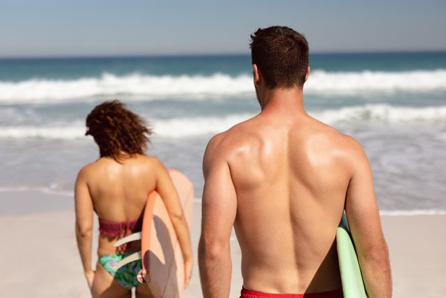 Rear view of diverse couple walking with surfboard on beach in the sunshine