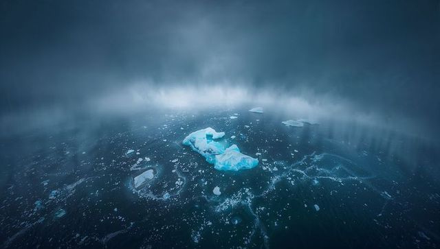 Luminescent iceberg fragment drifting through foggy arctic waters with scattered sea ice