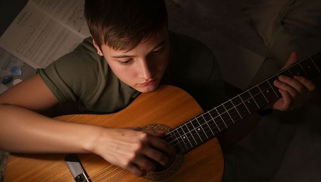 Teenager practicing acoustic guitar on bed with sheet music in low light, cozy intimate scene