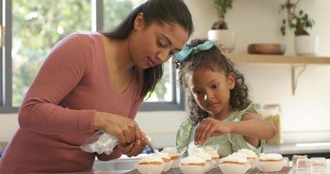 Mother teaching daughter cupcake decorating with piping bag and sprinkles in sunlit kitchen