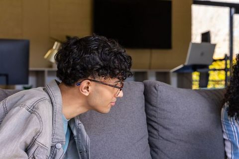 Young asian student leaning forward on gray sofa wearing glasses and denim jacket