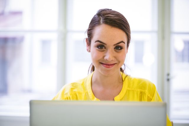 Young businesswoman smiling while working on laptop in a modern office. Ideal for use in business, technology, and career-related content, as well as advertisements and articles focusing on professional women and modern work environments.