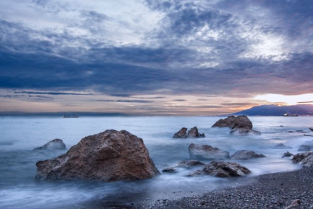 Image features a dramatic seaside landscape with large rocks on the shore under a cloudy sky at sunset. Ideal for use in travel brochures, nature-themed blogs, inspirational posters, and websites focused on outdoor adventures or wellness.