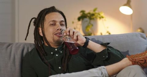 African american man reclining on couch drinking from glass bottle in cozy living room